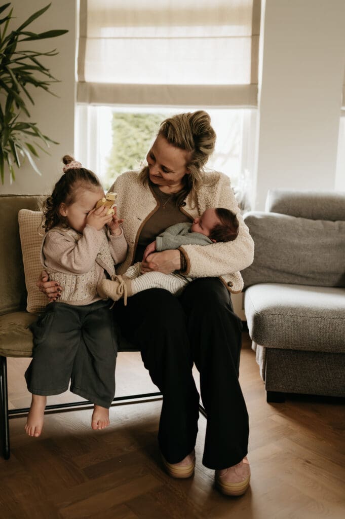 Ongedwongen lifestyle binnenshoot van een mama die genietend op de bank zit met baby en zusje.